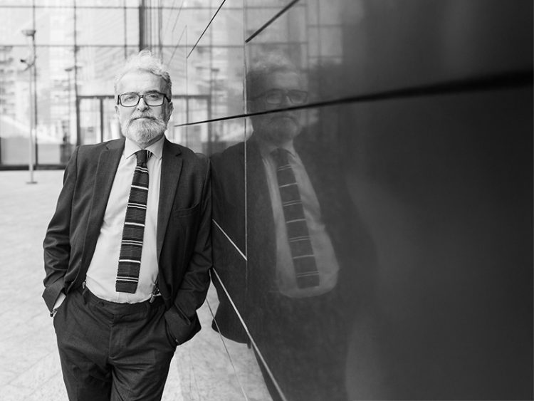 Senior businessman with hands in pockets leaning outside office building